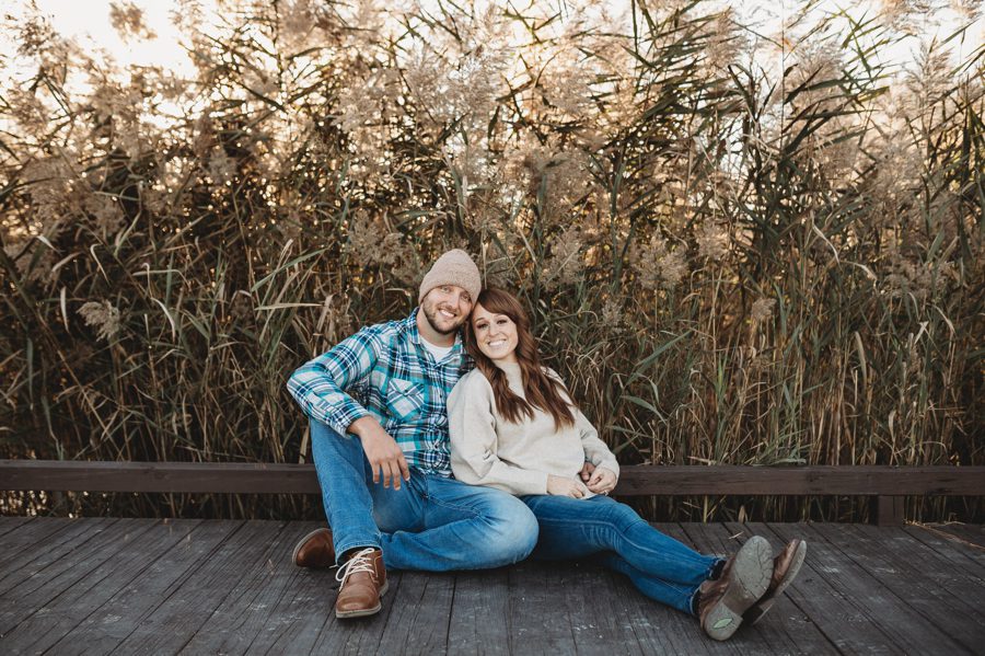 engaged couple wearing beanie with blue flannel and sweater at fall engagement while sitting on dock