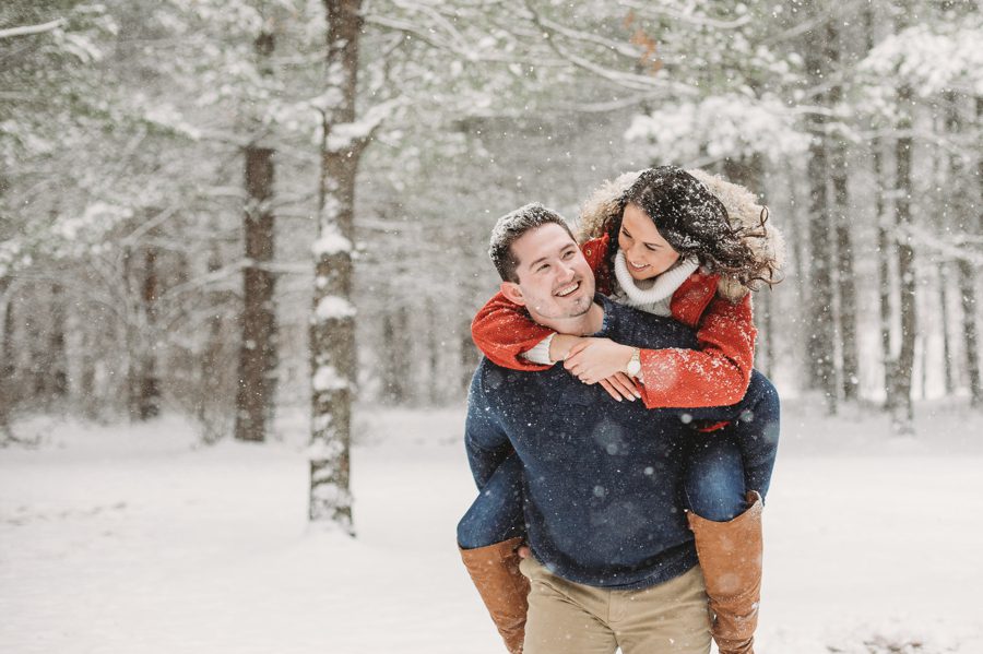 man giving fiancé in red coat a piggyback ride in the snow