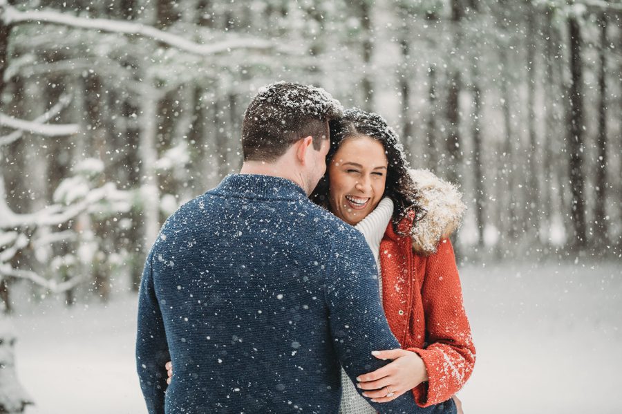 man in blue sweater whispering in fiancés ear wearing a red coat in the snow at Hoover Reservoir Engagement