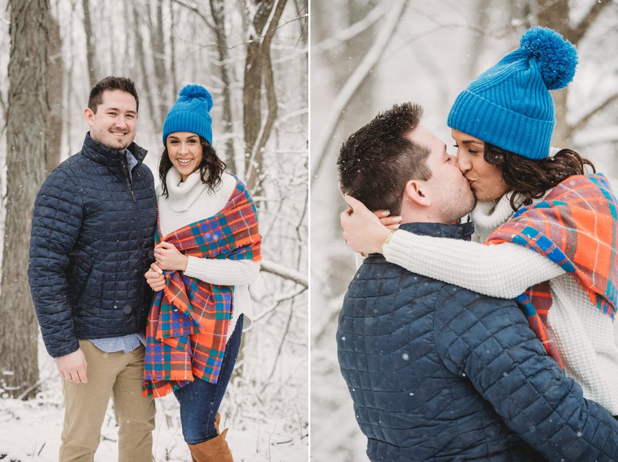 man in navy blue coat with fiancé in blue hat and plaid scarf in the snow with trees
