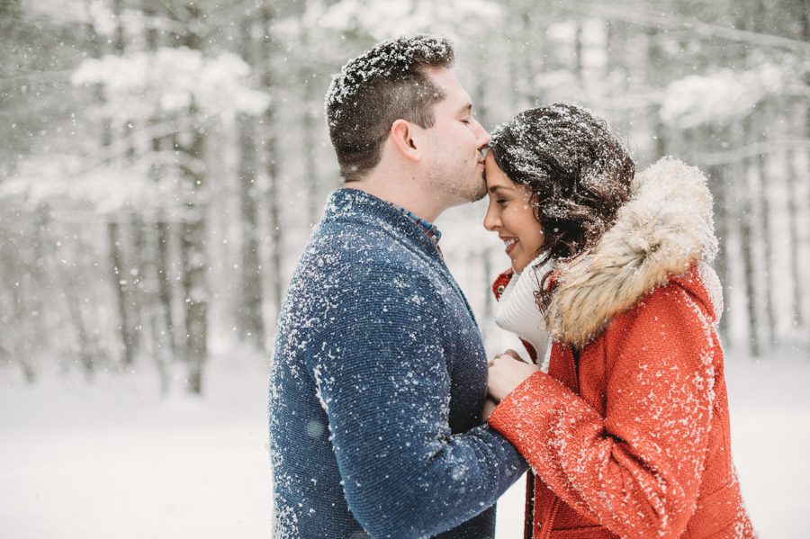groom to be kissing fiancés forehead in the snow with tall pines at Hoover Reservoir Engagement