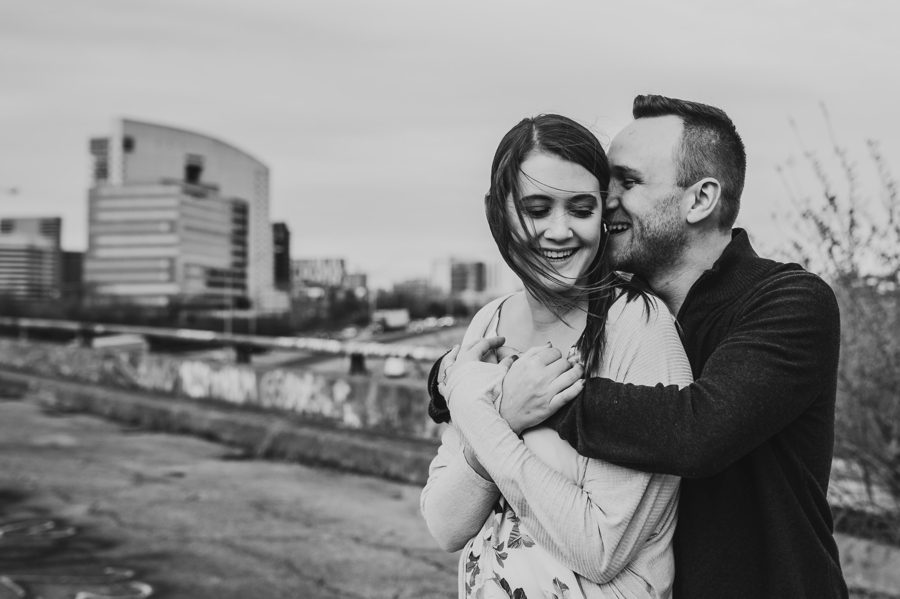 black and white image of man whispering in fiancés ear with her hair blowing
