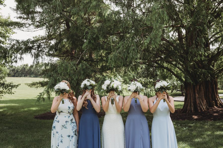 Bride with bridesmaids wearing mixed colored dresses holding white peony bouquets in front of their face